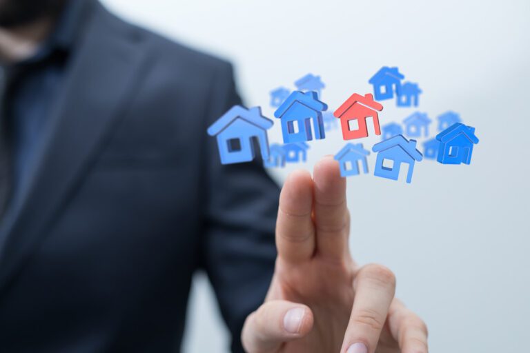 Casas em azul e uma em vermelha em primeiro plano. Esses ícones estão em cima da mão de uma pessoa.