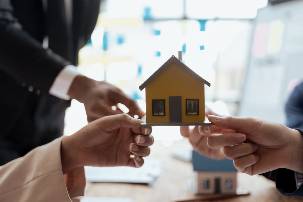 Mãos de uma equipe de vendas segurando um modelo de casa em miniatura, representando o planejamento estratégico para o mercado imobiliário.