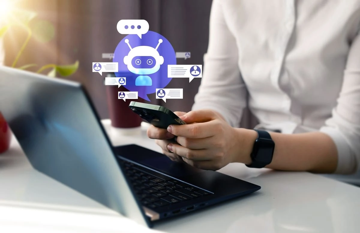 Pessoa utilizando smartphone e notebook, com representação gráfica de um chatbot com inteligência artificial e mensagens de atendimento ao redor, ilustrando o uso de chatbots para construtoras na comunicação e no relacionamento com clientes.